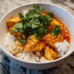 A close-up of a Spicy Coconut Curry Chicken Bowl featuring chunks of chicken in orange sauce over white rice, topped with fresh cilantro.