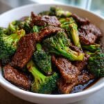 Close-up of tender beef slices coated in dark sauce mixed with bright green broccoli florets in a white bowl, showcasing Spicy Takeout Beef And Broccoli.