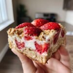 A hand holds a delicious slice of Strawberry Shortcake Baked Oats, topped with fresh strawberries and creamy filling.
