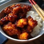 A bowl of fluffy white rice topped with glistening, crispy takeout orange chicken.