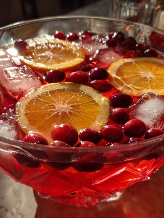 Close-up of a vibrant, red Thanksgiving Punch garnished with fresh cranberries, orange slices, and ice cubes in a crystal bowl.