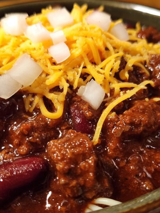 A close-up of a bowl of Ultimate Cincinnati Chili Recipe, topped with shredded cheddar cheese, diced onions, and kidney beans.