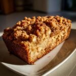 A golden-brown loaf of Vegan Apple Cinnamon Crumble Bread with a thick, crunchy topping, sitting on a white plate.