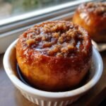 A close-up of a glossy Vegan Baked Cinnamon Caramel Apple topped with brown sugar crumble, served in a white ramekin.