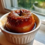 A single Vegan Baked Cinnamon Caramel Apple, glistening with glaze, served in a white ramekin by a sunny window.
