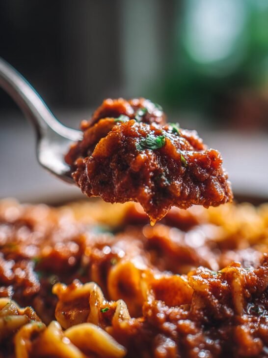 A spoonful of rich Vegan Bolognese sauce, garnished with herbs, lifted above a bowl of pasta.