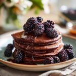 A stack of fluffy vegan chocolate pancakes topped with fresh blackberries and drizzled with chocolate syrup.
