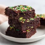 A stack of three rich, dark Vegan Fudge Zucchini Brownies topped with colorful sprinkles, showing green zucchini pieces inside.