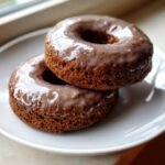 Two rich, dark Vegan Gingerbread Donuts Chai Glaze stacked on a white plate near a window.