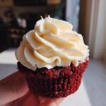 A close-up of a single Vegan Red Velvet Cupcakes topped with thick, swirled Bourbon Vanilla Buttercream frosting.