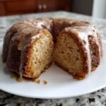 A close-up of an Apple Cider Donut Bundt Cake, sliced open to show the moist crumb and dripping with a light glaze.