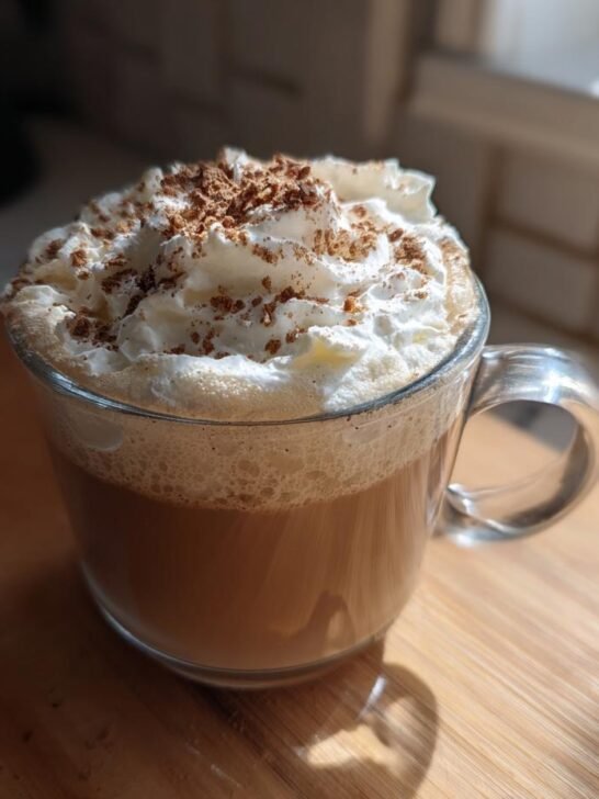 A glass mug filled with a creamy Apple Crisp Oatmilk Latte, topped high with whipped cream and cinnamon.
