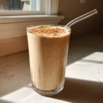 A tall glass filled with a creamy Apple Pie Smoothie, topped generously with ground cinnamon and a clear straw.