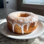 A single, perfectly baked vegan apple cider donut covered in thick white glaze, sitting on a small white plate.