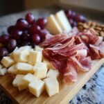 Close-up of cubed cheese, folded cured meats, red grapes, and almonds on a wooden board for How To Make The Best Charcuterie Board.