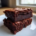 Close-up of two rich, dark, fudgy vegan brownies stacked on a white plate, showing a dense interior.