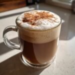 A rich Brown Butter Maple Cookie Latte topped with thick foam and sprinkled cinnamon in a clear glass mug.