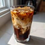 A tall glass filled with ice cubes, dark coffee, and cream swirling beautifully to make Brown Sugar Caramel Iced Coffee.