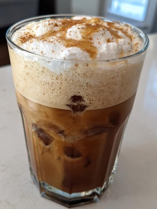 A tall glass filled with an iced Brown Sugar Iced Pumpkin Cream Latte, topped with foam, whipped cream, and cinnamon.