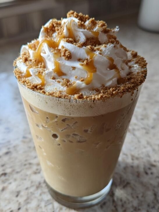A tall glass of Caramel Biscoff Cookie Butter Iced Latte topped with whipped cream, caramel drizzle, and cookie crumbs.