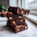 A stack of four rich, dark Chocolate Hazelnut Biscotti squares studded with visible hazelnuts on a white marble surface.