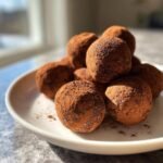 A close-up of several Chocolate Peanut Butter Energy Balls piled on a white plate, heavily dusted with cocoa powder.