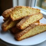 Close-up of several slices of golden Eggnog Biscotti Cookies stacked on a white plate near a window.