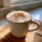 A close-up of steaming Eggnog White Hot Chocolate in a rustic white mug, topped with foam and cinnamon.
