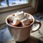 A creamy white mug filled with rich, dark Fancy Hot Chocolate With Marshmallows, sitting by a sunlit window.