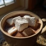 A close-up of a mug filled with rich, dark Fancy Hot Chocolate With Marshmallows, lit by sunlight.