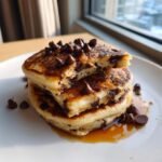 A stack of three fluffy vegan banana chocolate chip pancakes drizzled with syrup and topped with chocolate chips.