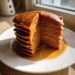 A tall stack of fluffy Vegan Gingerbread Pancakes cut to show the layers, drizzled heavily with syrup.