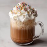 A clear glass mug filled with a layered Frosted Sugar Cookie Latte, topped with whipped cream and colorful sprinkles.