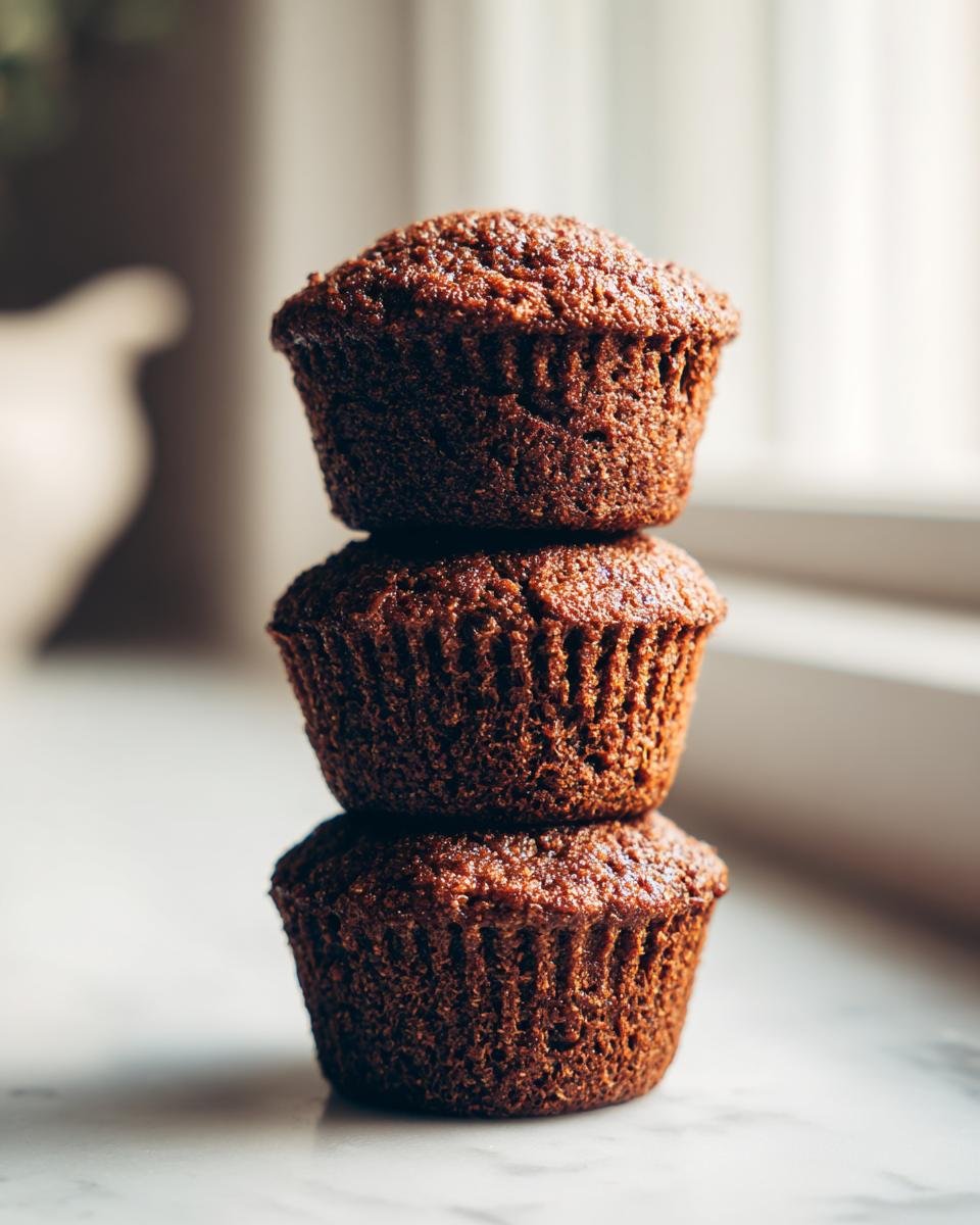 Three dark brown Healthy Vegan Gingerbread Quinoa Muffins stacked vertically on a light surface.