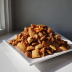 A large mound of glistening Homemade Everything Bagel Chex Mix featuring Chex cereal and pretzels, served on a white square plate.
