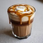Close-up of a layered Hot Honey Caramel Latte topped with foam and dripping caramel sauce.