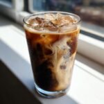 Close-up of a tall glass containing an Iced Brown Sugar Oat Milk Latte with swirling layers of coffee and oat milk over ice.