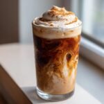 A tall, condensation-covered glass containing an iced Gingerbread Chai Tea Mocktail with swirling brown and white layers, topped with whipped cream and cinnamon.