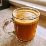 Close-up of a warm Maple Cinnamon Hot Buttered Rum Mocktail in a clear glass mug with a frothy top.