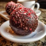 Close-up of a finished Peppermint Hot Cocoa Bombs, covered in dark chocolate and crushed candy canes, sitting on a white saucer.