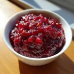 A close-up of The Perfect Apple Cranberry Sauce, rich red and chunky, topped with orange zest in a white bowl.