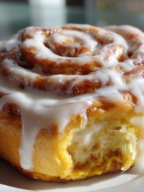 A single, freshly baked Pumpkin Cheesecake Cinnamon Roll generously topped with white cream cheese icing, sitting on a white plate.