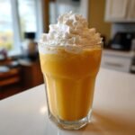 A tall glass filled with a thick, orange Pumpkin Cheesecake Milkshake, topped high with whipped cream.