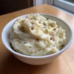 A white bowl filled with creamy Roasted Garlic Mashed Potatoes, topped with melted butter and herbs.
