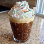 A tall glass of Starbucks Copycat Iced Sugar Cookie Latte topped with whipped cream and colorful sprinkles.