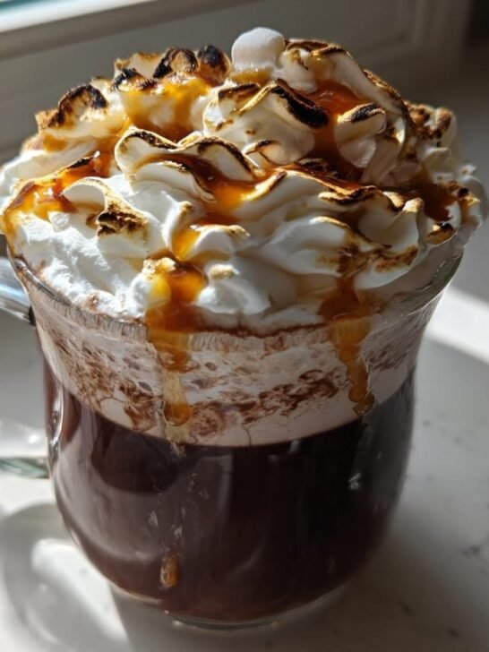 A clear glass mug filled with a dark Toasted Marshmallow Mocha topped with whipped cream, toasted marshmallows, and caramel drizzle.
