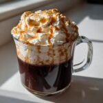 A rich Toasted Marshmallow Mocha topped with whipped cream, caramel drizzle, and toasted topping in a clear mug.