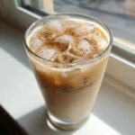 Close-up of a tall glass filled with a Vanilla Cinnamon Iced Latte, ice cubes, and a dusting of cinnamon.