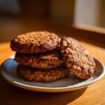 A stack of four golden-brown Vegan Chai Spiced Oatmeal Cookies on a small plate, with one cookie broken open.