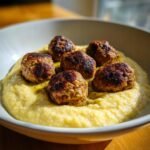 Six browned Vegan Chickpea Quinoa Meatballs served atop a bed of creamy yellow polenta in a white bowl.